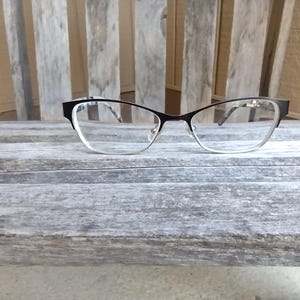 Może przedstawiać: Czarne okulary w stylu „kocie oko” z przezroczystymi szkłami. Okulary leżą na postarzanej drewnianej powierzchni.