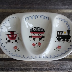 Puede incluir: Un plato de servir de cerámica blanca con tres secciones, cada una con un diseño de tren diferente. La sección central tiene un vagón de tren rojo con ruedas negras y un vagón de carbón negro. La sección izquierda tiene un vagón de tren rojo con ruedas negras. La sección derecha tiene una locomotora negra con una luz amarilla y ruedas negras. Cada sección tiene un borde azul y blanco festoneado.