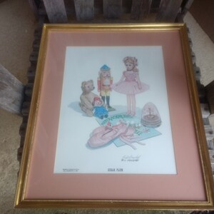 Könnte beinhalten: Gerahmter Druck mit einer Ballerina, einem Nussknacker, einem Teddybären und Spielzeug. Das Kunstwerk hat ein rosa-goldenes Farbschema, mit Ballettschuhen im Vordergrund. Der Druck befindet sich in einem goldenen Rahmen.