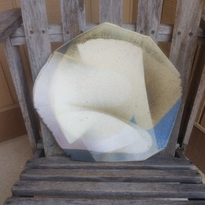 Może przedstawiać: Biały ceramiczny talerz z niebieską, brązową i fioletową glazurą. Talerz ma nieregularny kształt i stoi na drewnianym krześle.