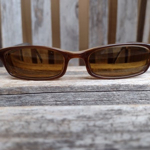 Może przedstawiać: Okulary przeciwsłoneczne z brązową plastikową oprawką i brązowymi szkłami. Okulary leżą na drewnianej powierzchni.