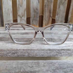 Può includere: Occhiali con montatura trasparente a forma di occhi di gatto. Gli occhiali hanno una tinta marrone chiaro e sono appoggiati su uno sfondo di legno stagionato. Le montature sono in materiale trasparente.