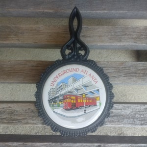 May include: Black cast iron hot pad with a white ceramic tile featuring a red and white train in front of a building with the text "UNDERGROUND ATLANTA".