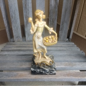 Può includere: Una statuetta di una donna che tiene un cesto di frutta. La donna indossa un vestito bianco con una fascia blu e un corpetto giallo. Il cesto è fatto di materiale intrecciato ed è pieno di frutta gialla e arancione. La statuetta è in piedi su una base di marmo nero.