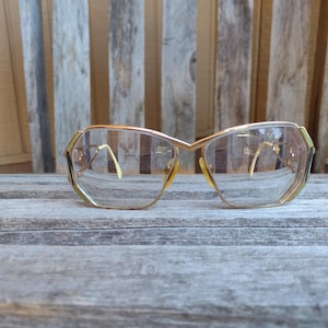 Peut inclure: Lunettes vintage avec une monture en métal doré et des verres transparents. Les lunettes ont une forme géométrique unique avec un subtil accent vert sur les coins. Les branches sont de couleur jaune clair.