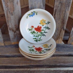 May include: A stack of four small, off-white ceramic bowls with a gold rim. Each bowl features a floral design with red, yellow, and blue flowers. The bowls are stacked on a wooden surface.