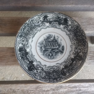 Puede incluir: Plato de porcelana blanco y negro con un borde decorativo y una imagen central de un edificio con un jardín. El plato tiene un borde dorado.