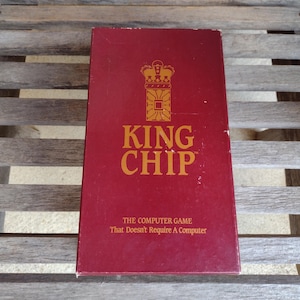 Op de afbeelding: Een rechthoekige, bordeauxrode doos voor het spel "King Chip" met gouden letters en een kroonembleem. De tekst luidt "THE COMPUTER GAME That Doesn't Require A Computer". De doos ligt op een houten lattenoppervlak.