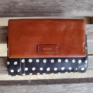 May include: A brown leather Fossil wallet with a black and white polka dot pattern. The wallet has a rectangular shape with a flap closure and the Fossil logo embossed on the front.
