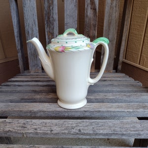 Può includere: Una caffettiera in ceramica bianca con un design floreale verde. La caffettiera ha un coperchio con un pomello verde e un manico con un accento verde.