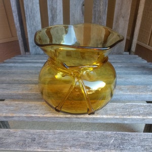 Può includere: Vaso in vetro ambrato con un designo annodato, che ricorda una borsa. Il vaso ha un'ampia apertura e un corpo arrotondato.