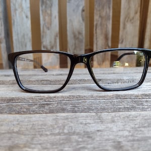May include: Black rectangular eyeglasses with clear lenses. The frames have a thick, bold design. The brand name "RALPH LAUREN" is printed on the lower right lens. The glasses are set against a wooden background.