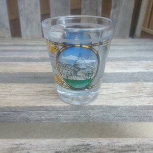 May include: A clear glass shot glass with a gold rim and a colorful image of the United States Capitol building. The image is surrounded by a gold frame and includes the words "Washington D.C."