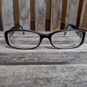 Może przedstawiać: Czarne okulary z prostokątną oprawą i przezroczystymi soczewkami. Okulary leżą na tle zwietrzałego drewna. Oprawki mają subtelny, zaokrąglony kształt, a zauszniki są cienkie i eleganckie.