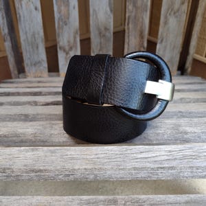 May include: A black leather belt with a textured surface and a large, rounded buckle. The buckle has a silver-colored metal finish. The belt is resting on a weathered wooden surface, suggesting a rustic or vintage aesthetic.