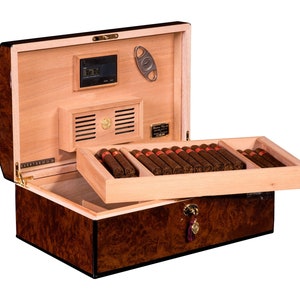May include: A brown wooden humidor with a hinged lid, open to reveal a cedar-lined interior with a built-in humidifier and a cigar cutter. The humidor is filled with rows of brown cigars.
