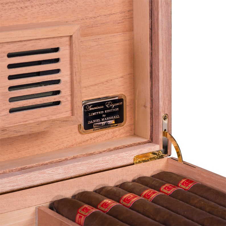 May include: A wooden cigar box with a gold-coloured metal plaque that reads "American Elegance Limited Edition by Daniel Marshall No. 35/200". The box is open and contains several cigars.