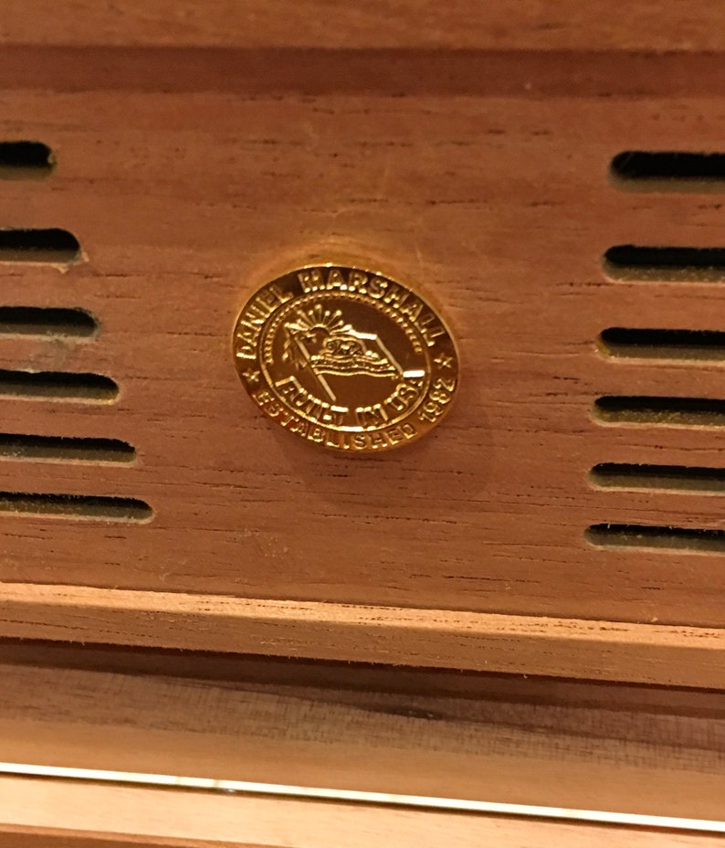 May include: A gold-coloured metal emblem with the text "Daniel Marshall * Family * Established 1982 * NY USA" on a wooden surface.