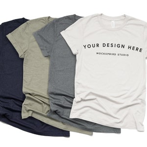 Puede incluir: Cuatro camisetas de diferentes colores, incluyendo azul marino, verde oliva, gris jaspeado y blanco. La camiseta blanca tiene el texto "YOUR DESIGN HERE" y "MOCKUPBIRD STUDIO" impreso en ella.