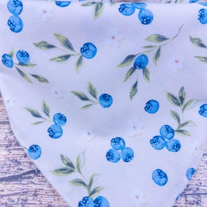 May include: A light-colored fabric with a repeating pattern of blueberries and leaves. The blueberries are a vibrant blue, and the leaves are green. Small white flowers are also scattered throughout the design. The fabric is folded, creating a triangular shape.