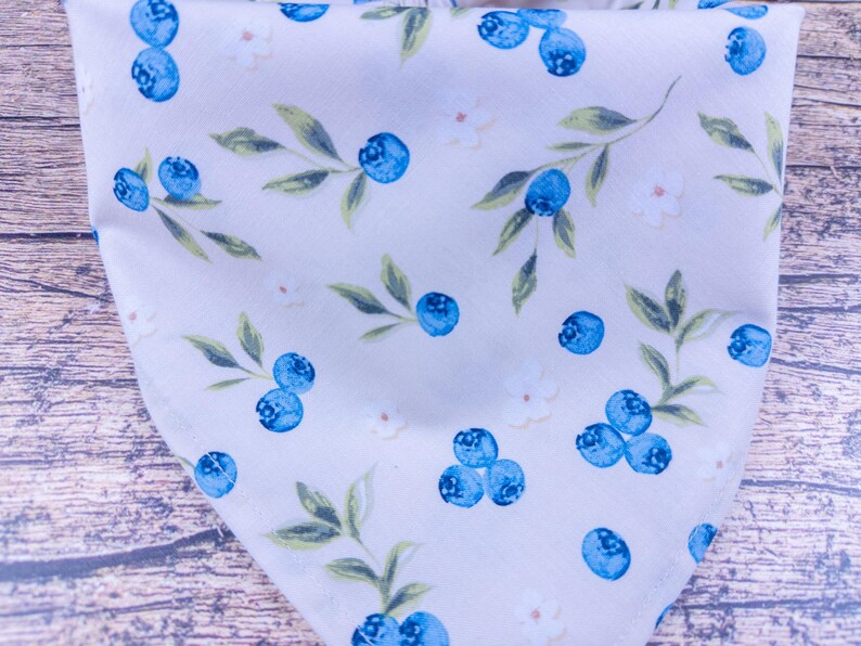 May include: A light-colored fabric with a repeating pattern of blueberries and leaves. The blueberries are a vibrant blue, and the leaves are green. Small white flowers are also scattered throughout the design. The fabric is folded, creating a triangular shape.