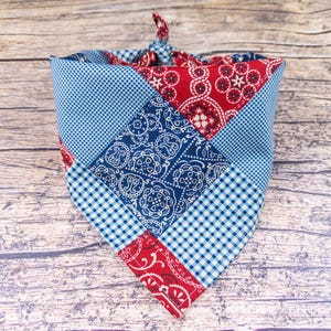 May include: A patchwork dog bandana in red, white, and blue. The bandana features a mix of patterns, including paisley, gingham, and polka dots. The bandana is tied at the top, ready to be worn.