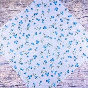 May include: White fabric with a pattern of blue blueberries and green leaves. The fabric is arranged diagonally on a wooden surface. Small white flowers are also scattered throughout the design.