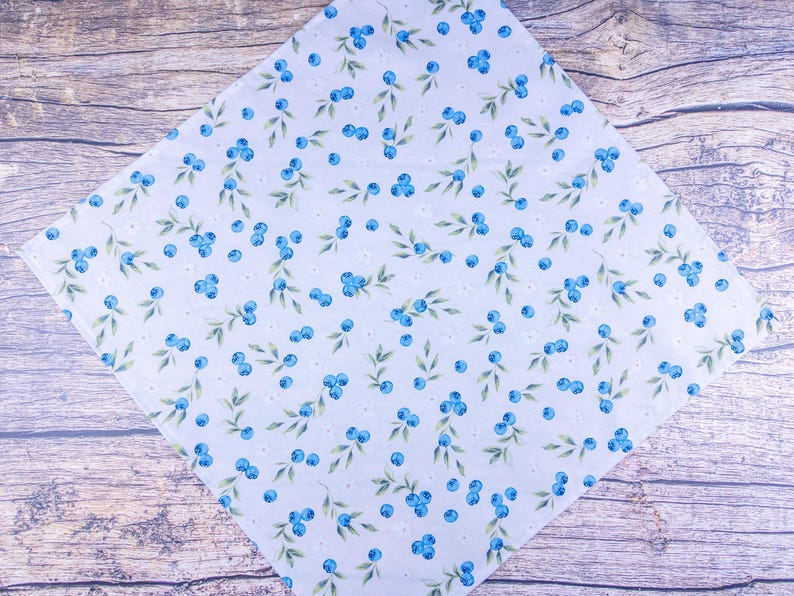 May include: White fabric with a pattern of blue blueberries and green leaves. The fabric is arranged diagonally on a wooden surface. Small white flowers are also scattered throughout the design.