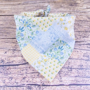 May include: A floral patterned dog bandana with a light blue, yellow, and white color scheme. The bandana features a patchwork design with floral and gingham patterns. The top is tied in a knot.