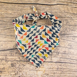 May include: A colorful bandana with a geometric pattern of triangles in shades of blue, red, yellow, and orange on a white background. The bandana is tied in a knot at the top.