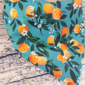 May include: A teal blue bandana with a repeating pattern of orange citrus fruit and white flowers. The bandana is made of fabric and has a stitched edge.