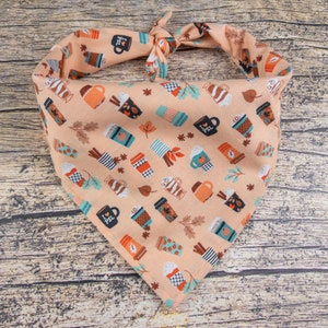 May include: A peach-colored bandana with a pattern of coffee cups and fall leaves. The bandana is tied in a knot at the top.