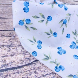 May include: A white bandana with a pattern of blue blueberries, green leaves, and small white flowers. The fabric is folded, showing the pattern at different angles. The bandana is likely made of soft cotton or a similar material.