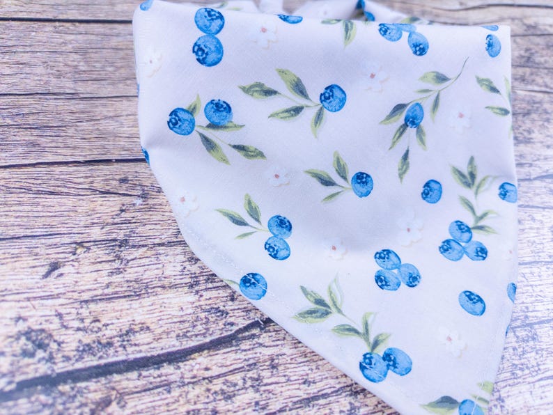 May include: A white bandana with a pattern of blue blueberries, green leaves, and small white flowers. The fabric is folded, showing the pattern at different angles. The bandana is likely made of soft cotton or a similar material.
