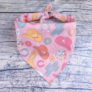 May include: Pink bandana with a western theme featuring cacti, cowboy hats, boots, and horseshoes. The bandana is tied in a knot at the top.