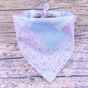 May include: A patchwork bandana in shades of lavender, white, and light blue. The fabric features floral and gingham patterns. The bandana is knotted at the top, creating a triangular shape.