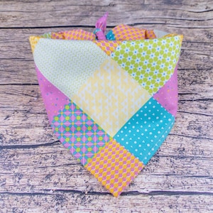 May include: A colorful patchwork dog bandana with a variety of patterns, including polka dots, floral, and geometric designs. The bandana features a mix of pink, yellow, green, and blue fabrics, tied at the top with a pink knot.