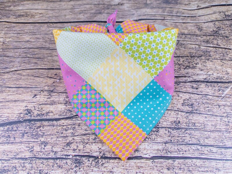 May include: A colorful patchwork dog bandana with a variety of patterns, including polka dots, floral, and geometric designs. The bandana features a mix of pink, yellow, green, and blue fabrics, tied at the top with a pink knot.