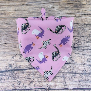 May include: A lavender dog bandana with a Halloween dinosaur theme. The bandana features various dinosaur illustrations, including skeleton, zebra-striped, and purple dinosaurs, along with jack-o'-lanterns. The bandana is tied at the top.