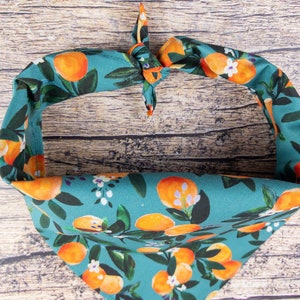 May include: A teal blue bandana with a pattern of orange citrus fruit and white flowers. The bandana is tied in a knot at the top.