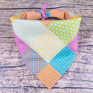 May include: A colorful patchwork dog bandana with a variety of patterns, including polka dots, floral designs, and geometric shapes. The bandana features a mix of pink, yellow, blue, and green fabrics, tied at the top.