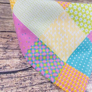 May include: A colorful patchwork fabric item with various patterns and colors. The design includes squares of pink with blue dots, yellow with a small pattern, and light blue with white dots. The item is on a wooden surface.