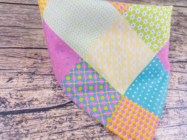 May include: A colorful patchwork fabric item with various patterns and colors. The design includes squares of pink with blue dots, yellow with a small pattern, and light blue with white dots. The item is on a wooden surface.