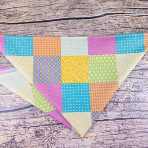 May include: A colorful, patchwork-style dog bandana in a triangular shape. The bandana features squares of various patterns and colors, including blue with white dots, yellow with orange dots, and pink with purple diamonds. The background is a wooden surface.