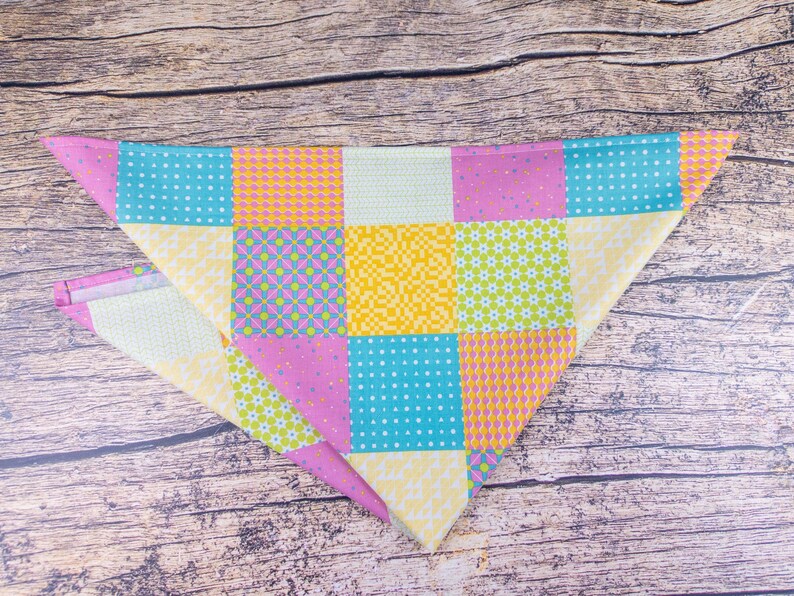May include: A colorful, patchwork-style dog bandana in a triangular shape. The bandana features squares of various patterns and colors, including blue with white dots, yellow with orange dots, and pink with purple diamonds. The background is a wooden surface.