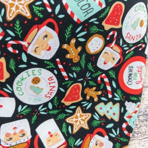 May include: Black fabric with a repeating pattern of Christmas-themed illustrations. The pattern includes gingerbread men, candy canes, cookies, and mugs with Santa's face. The text "Cookies for Santa" is repeated throughout the pattern.