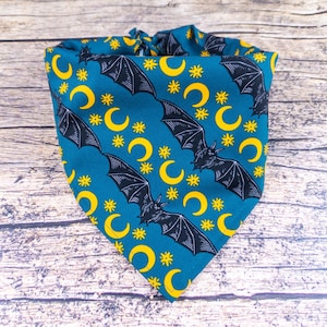 May include: A teal pet bandana with a repeating pattern of black bats, yellow crescent moons, and yellow stars. The bandana is tied at the top and is made of fabric. The background is a wooden surface.