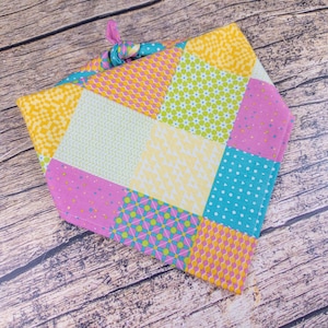 May include: A colorful patchwork bandana featuring squares of yellow, pink, turquoise, and green fabric. The bandana has various patterns, including polka dots, floral, and geometric designs. A knotted tie secures the bandana.