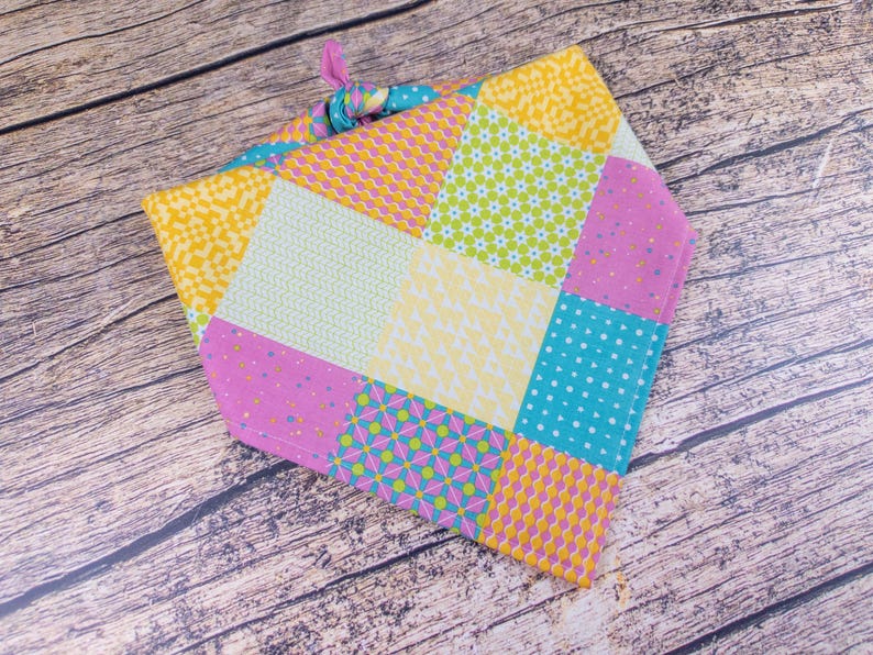 May include: A colorful patchwork bandana featuring squares of yellow, pink, turquoise, and green fabric. The bandana has various patterns, including polka dots, floral, and geometric designs. A knotted tie secures the bandana.