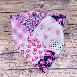 May include: A patchwork dog bandana featuring various floral and patterned fabrics in pink, white, and navy blue. The bandana is tied at the top, showcasing a mix of gingham, floral, and polka dot designs. The overall aesthetic is feminine and handcrafted.